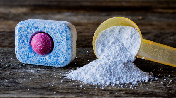 Dishwasher tablet and yellow measuring spoon with dishwashing powder on wooden surface.