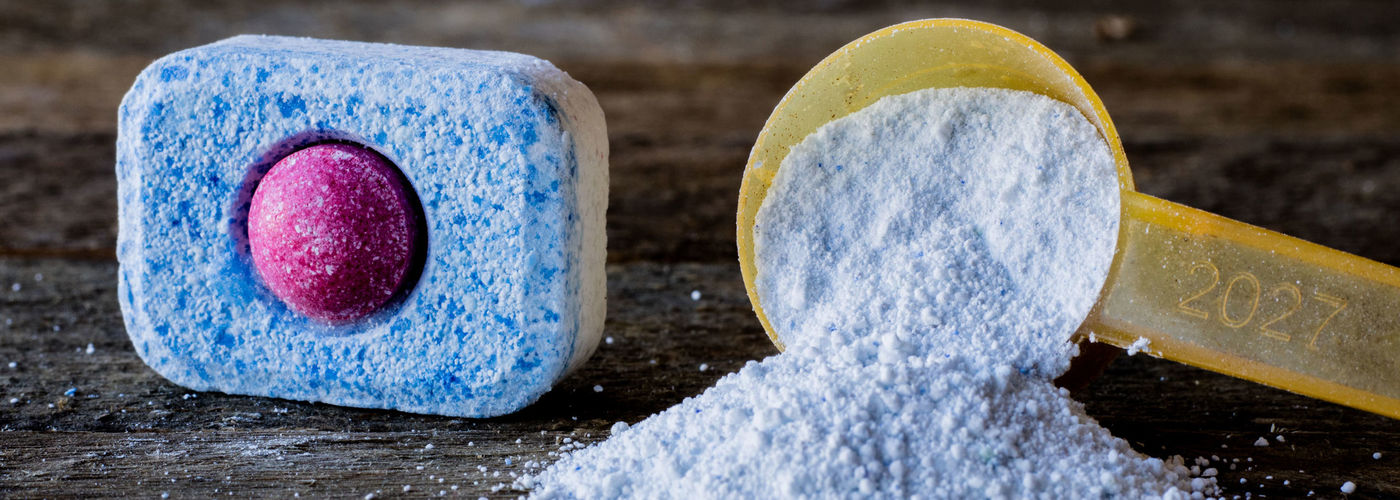 Dishwasher tablet and yellow measuring spoon with dishwashing powder on wooden surface.