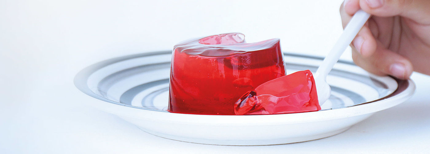 red gelatine dessert portion on a plate with a striped pattern, a piece of which is lifted out with a white spoon.