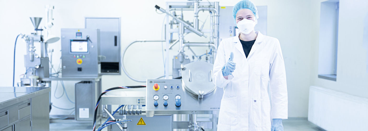 Person in protective clothing and lab coat stands in front of a machine in a sterile laboratory and gives a thumbs up.